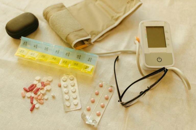 Flat lay image of healthcare essentials including a blood pressure monitor, pills, and eyeglasses on a white surface.