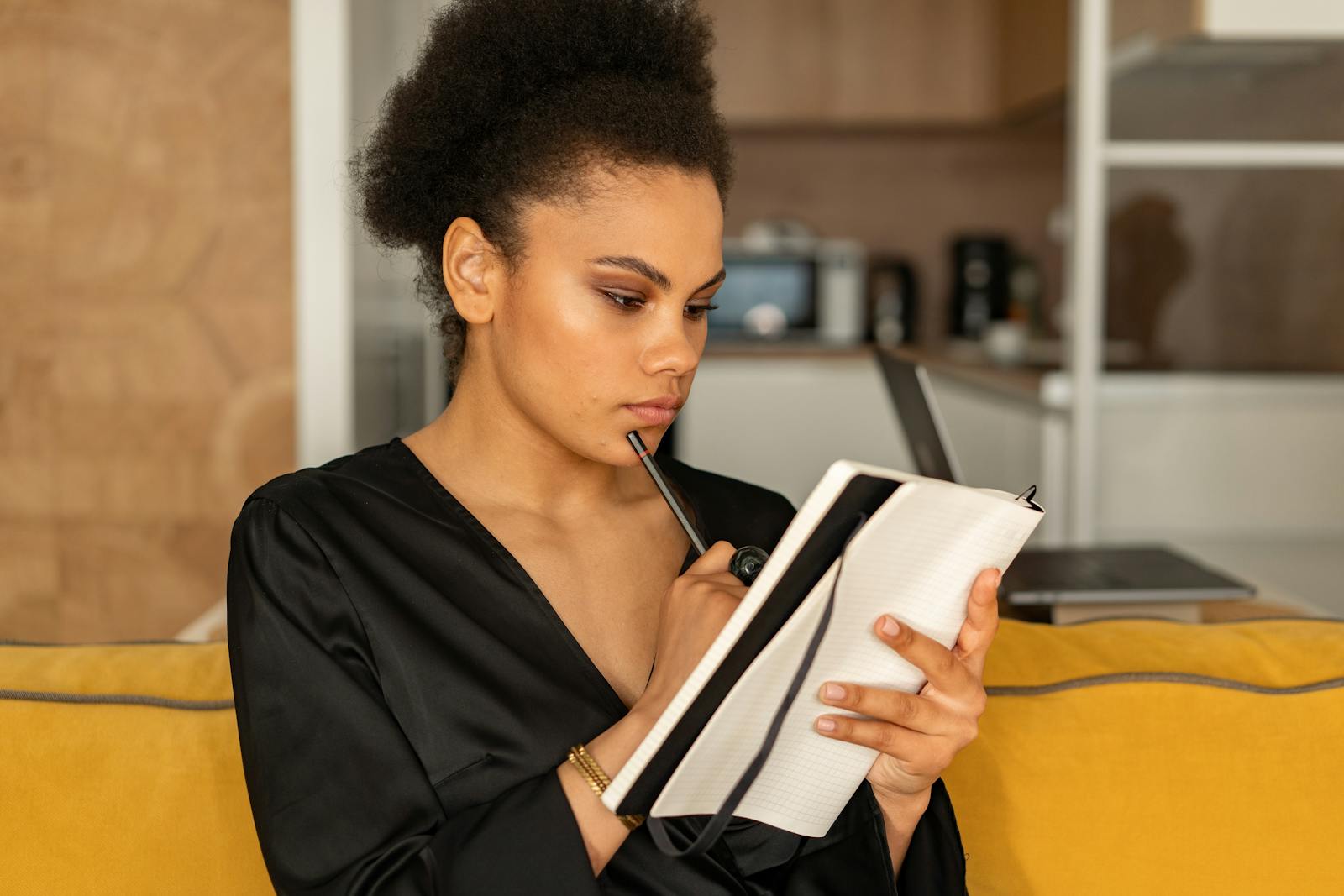 Focused woman in black attire pens thoughts in a notebook while sitting on a cozy sofa at home.